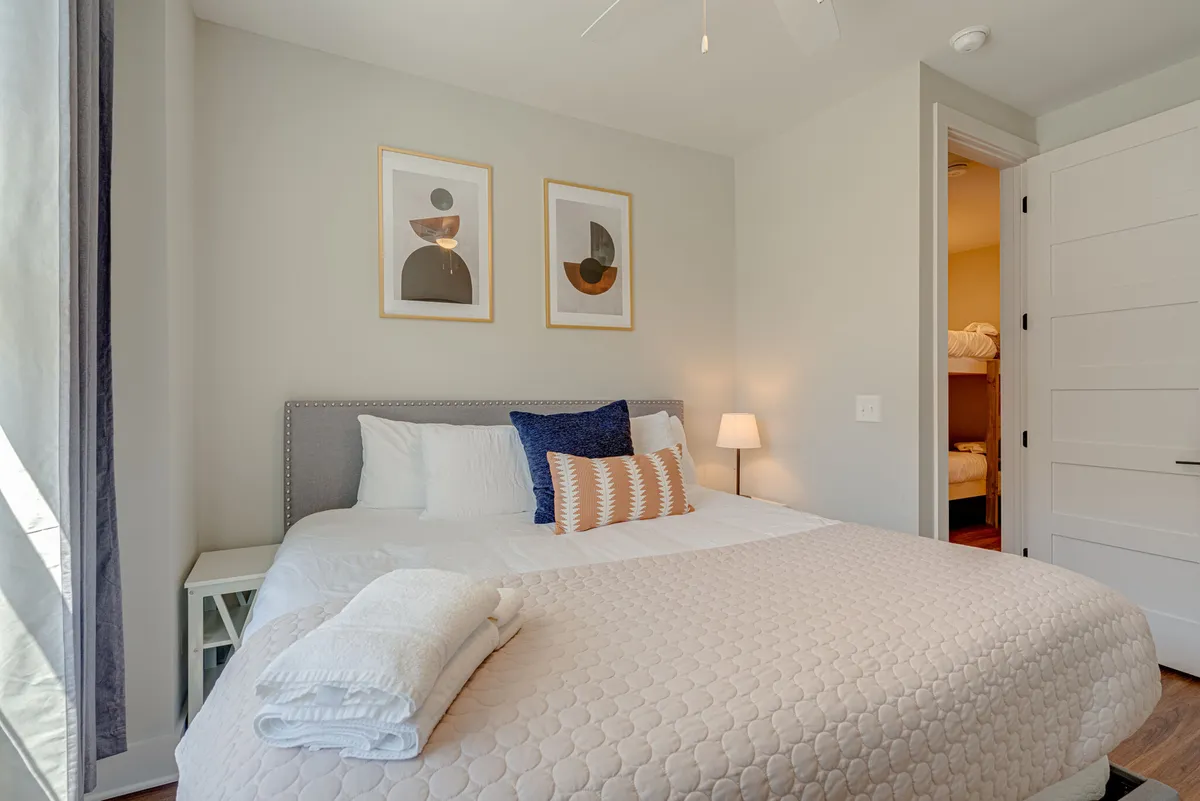 A serene bedroom with a king-sized bed adorned with decorative pillows, a nightstand with a lamp, and framed artwork. Gentle natural light filters through, creating a calm and inviting atmosphere ideal for rest.