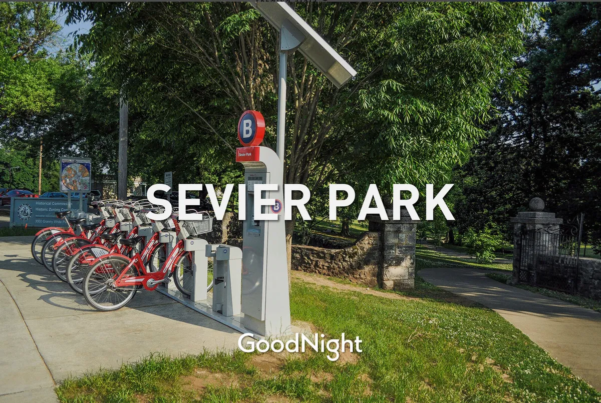A lively park entrance with a bicycle rental station, park sign, and inviting green spaces, suggesting opportunities for active outings like biking, walking, or picnicking. The scene is community-oriented and family-friendly, with a focus on outdoor recreation.