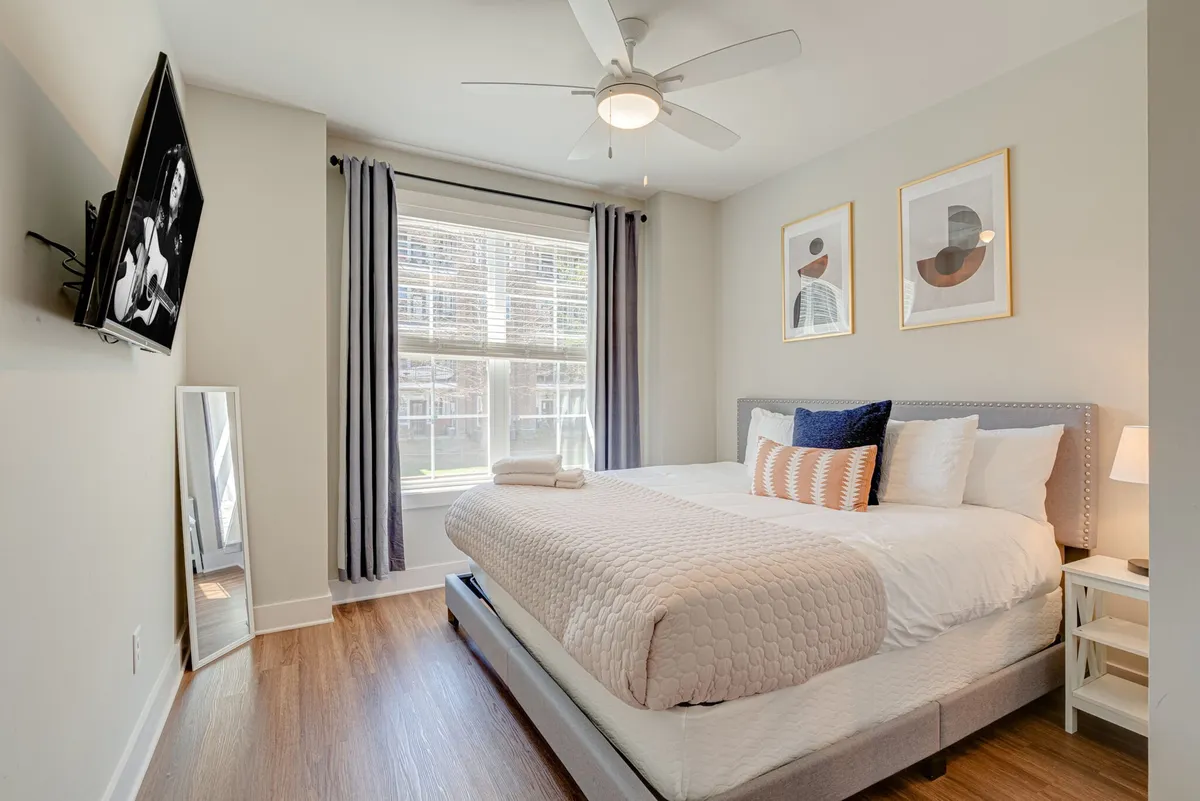 A sunlit bedroom with a king-sized bed, soft white linens, a large window letting in natural light, modern artwork above the bed, a stylish mirror, and a Smart TV mounted on the wall. The overall look is relaxed and cozy, evoking a sense of tranquility and comfort.