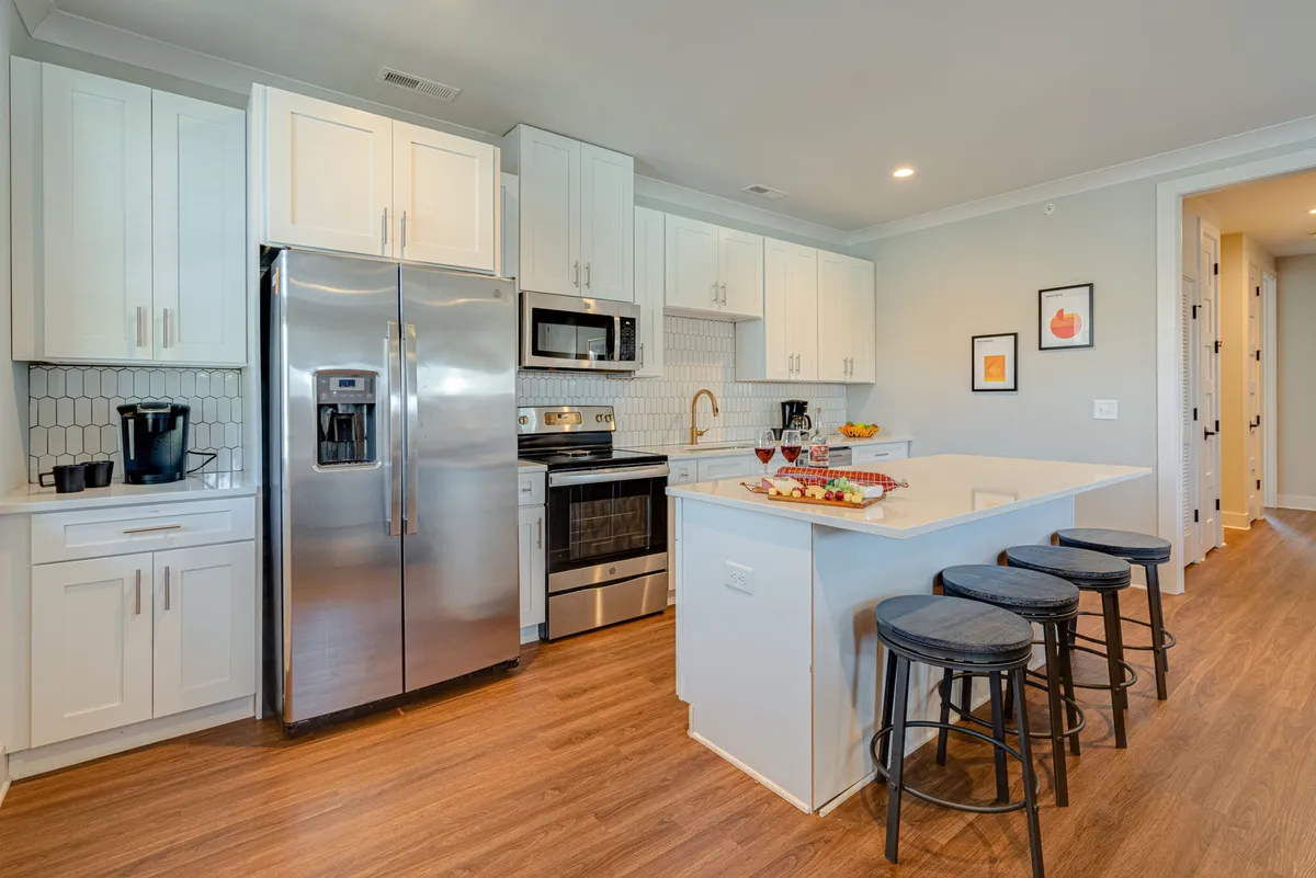 A bright, modern kitchen featuring stainless steel appliances, crisp white cabinetry, a central island with bar stools, and a colorful fruit display. Decorative wall art on the side adds personality, while the open design allows for easy movement and group cooking.