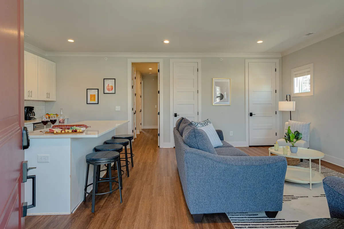 An open-concept living area that seamlessly connects the lounge, kitchen island with bar stools, and dining space. The sofa and coffee table are arranged for conversation, and wall art adds character. The overall design is modern, warm, and perfectly suited for hosting or relaxing.